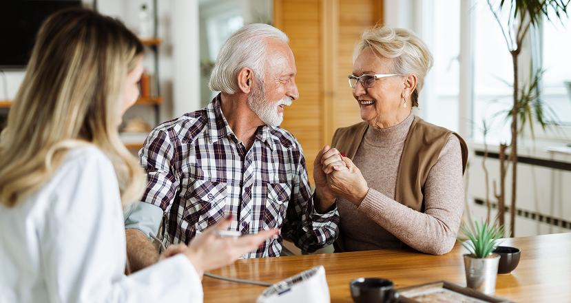 elderly couple at home holding hands and smiling with their doctor